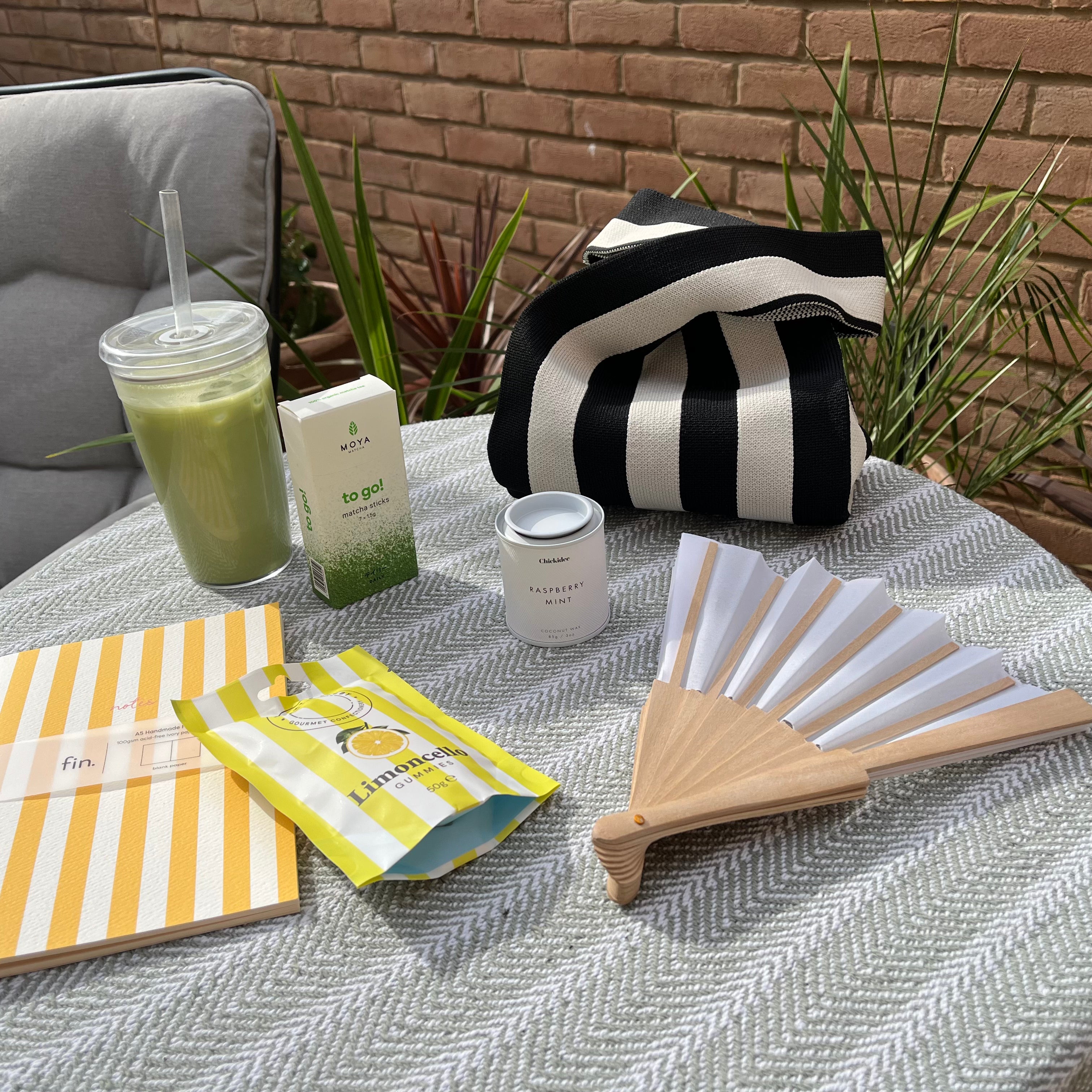 A styled outdoor table scene with multiple products: a black-and-white striped tote bag, a yellow-and-white striped notebook, a yellow and blue packaged item, a small white tin, a green drink, and a folding fan, arranged on a gray herringbone tablecloth with plants and a brick wall in the background.