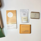 Five coffee-related items arranged on a white surface: a cream and tan card reading 'Aromatic of the Citric Moon' with a circular moon graphic, a white pouch labeled 'Vanilla' with 'Earthbound Coffee Roasts' and '100% Arabica', a small square box labeled 'WS + Co' and 'Handcrafted Coffee', a tan card reading 'Secret Salt Society' and 'Bergamot Pomegranate & Orange Blossom', and a tan rectangular item with 'CITRUS' printed on it.