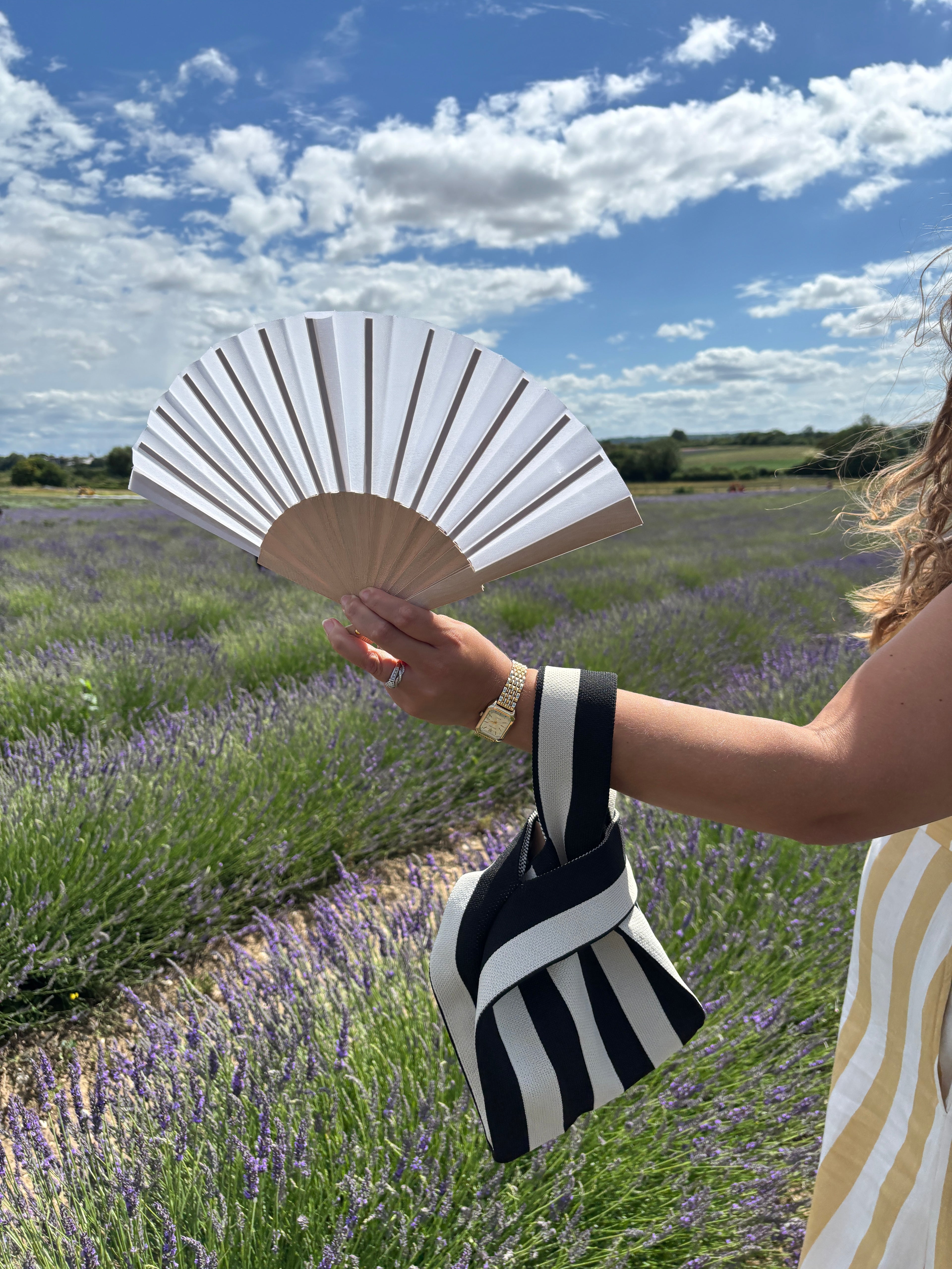 A person outdoors in a lavender field holding a black and white striped folding fan and matching striped bag, with a blue sky and clouds in the background.