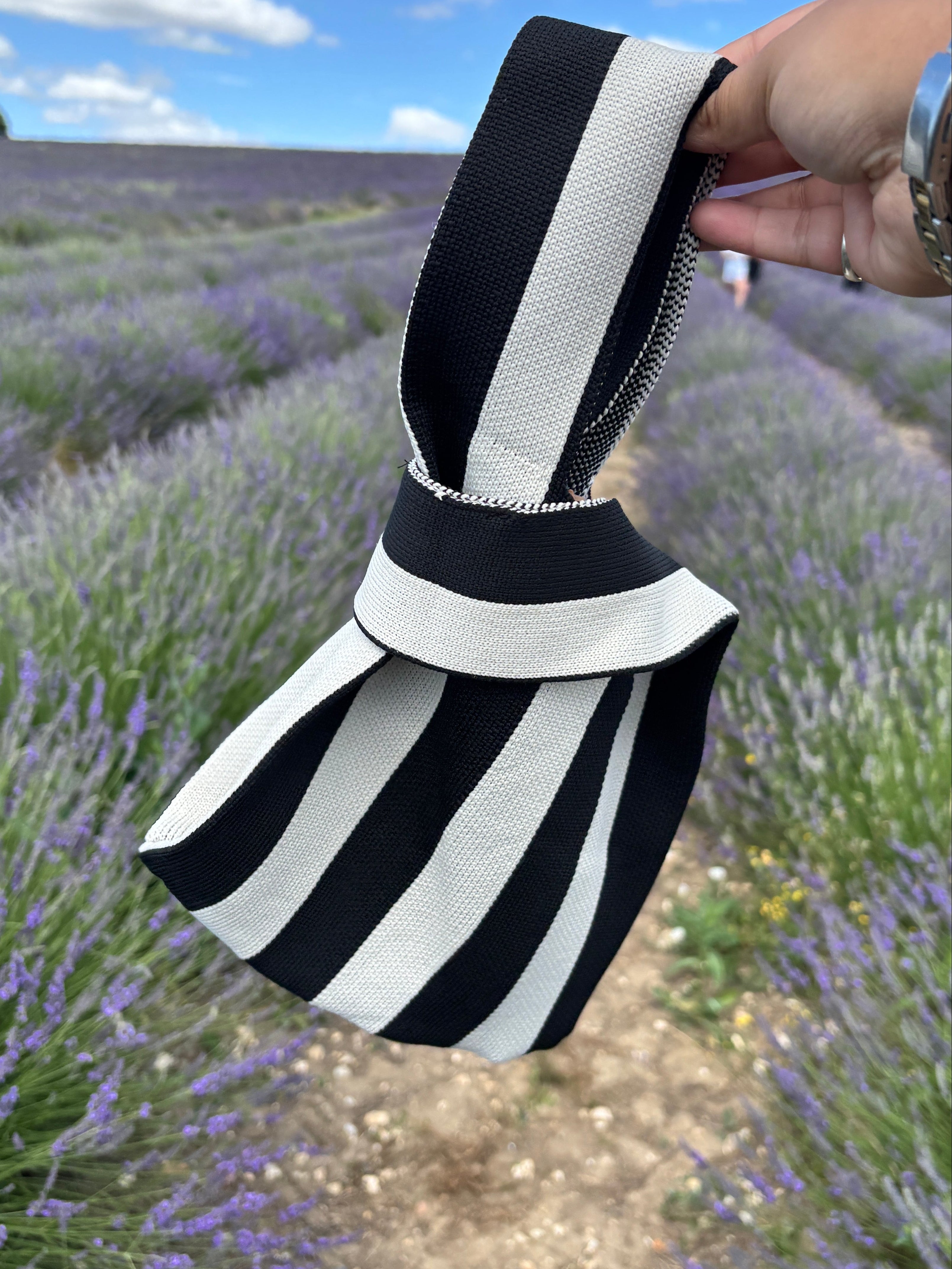 A hand holding a black and white striped fabric bag with a zipper closure, set against a background of a lavender field under a blue sky.