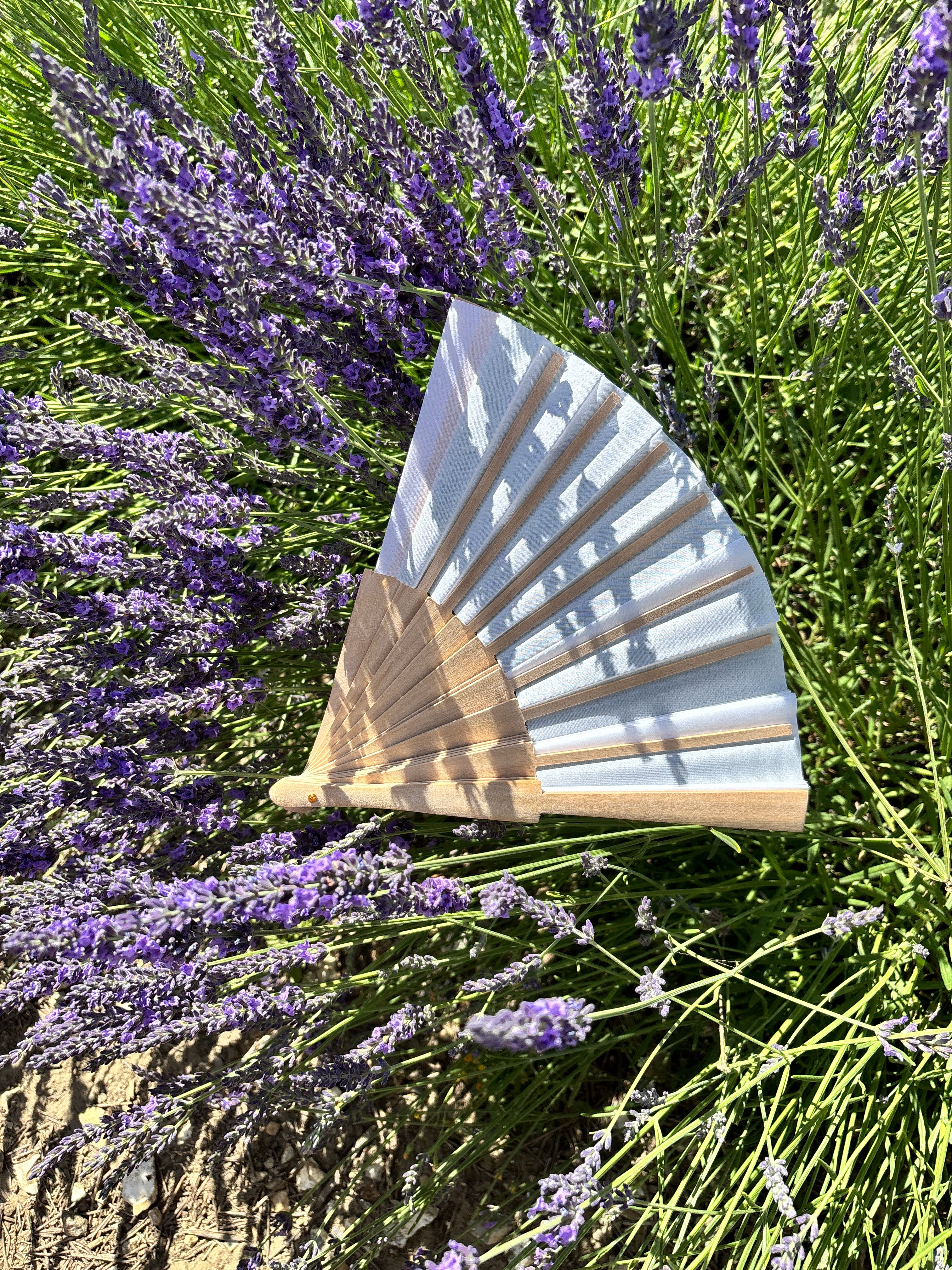 A folding hand fan with a white fabric upper section and a natural wood lower section, displayed against a background of green stems and purple lavender flowers.